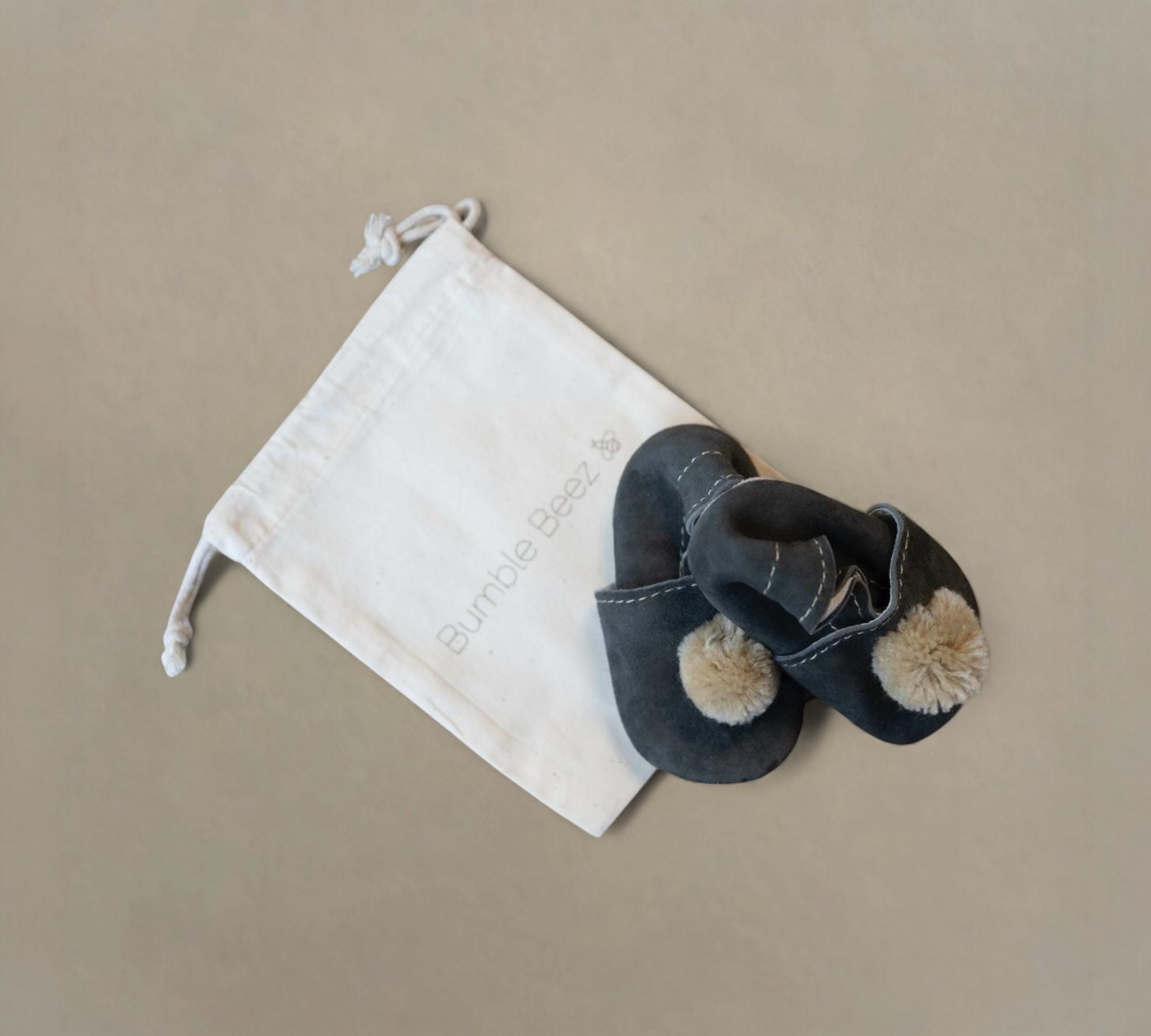Handcrafted genuine leather and suede Pom Pom Baby Slippers in dark color with elastic back, presented with a cotton drawstring bag