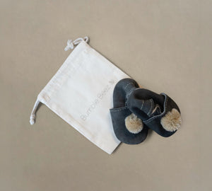 Handcrafted genuine leather and suede Pom Pom Baby Slippers in dark color with elastic back, presented with a cotton drawstring bag