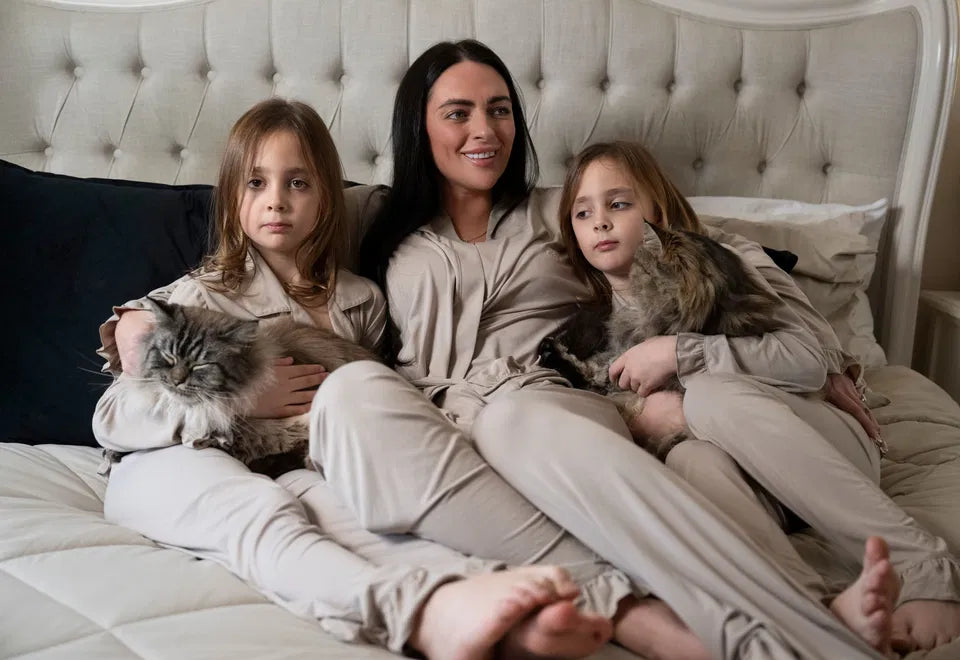 Mother and two children wearing matching Cinereous Luxe Lounge Sets in warm beige taupe, sitting on bed holding cats.