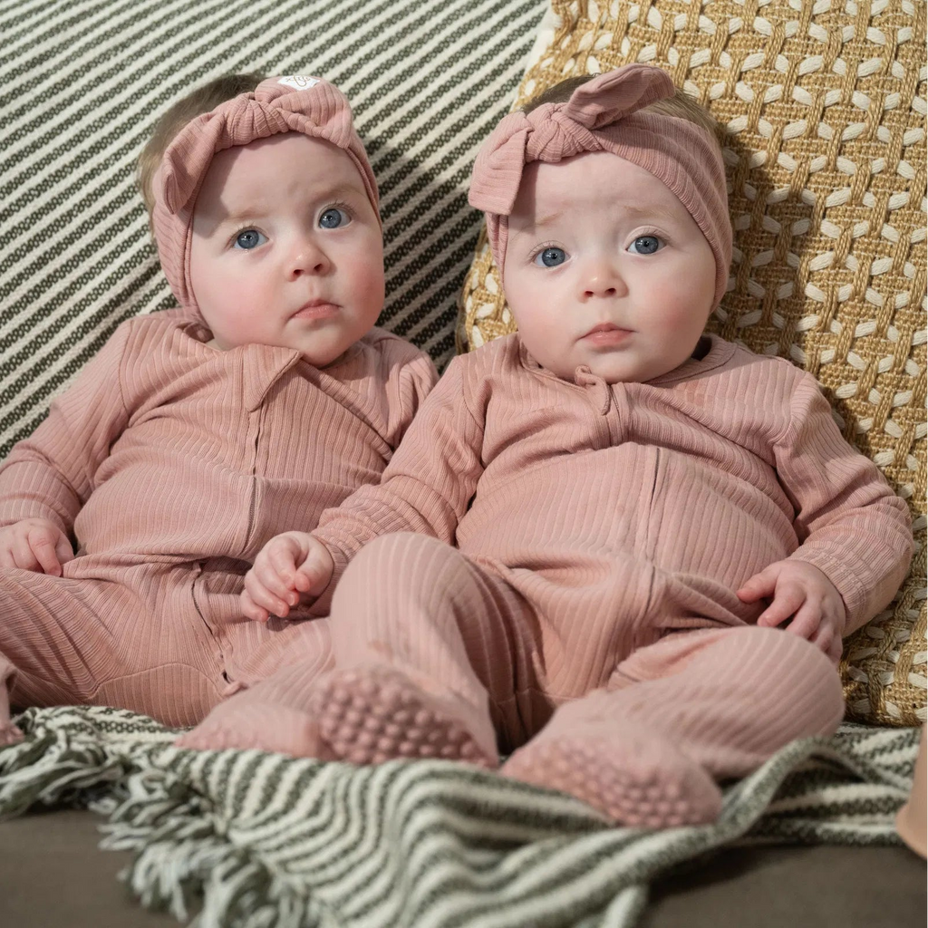 Two babies wearing The Ribbed Set Bow Edition in rose pink ribbed cotton sitting on striped and woven cushions