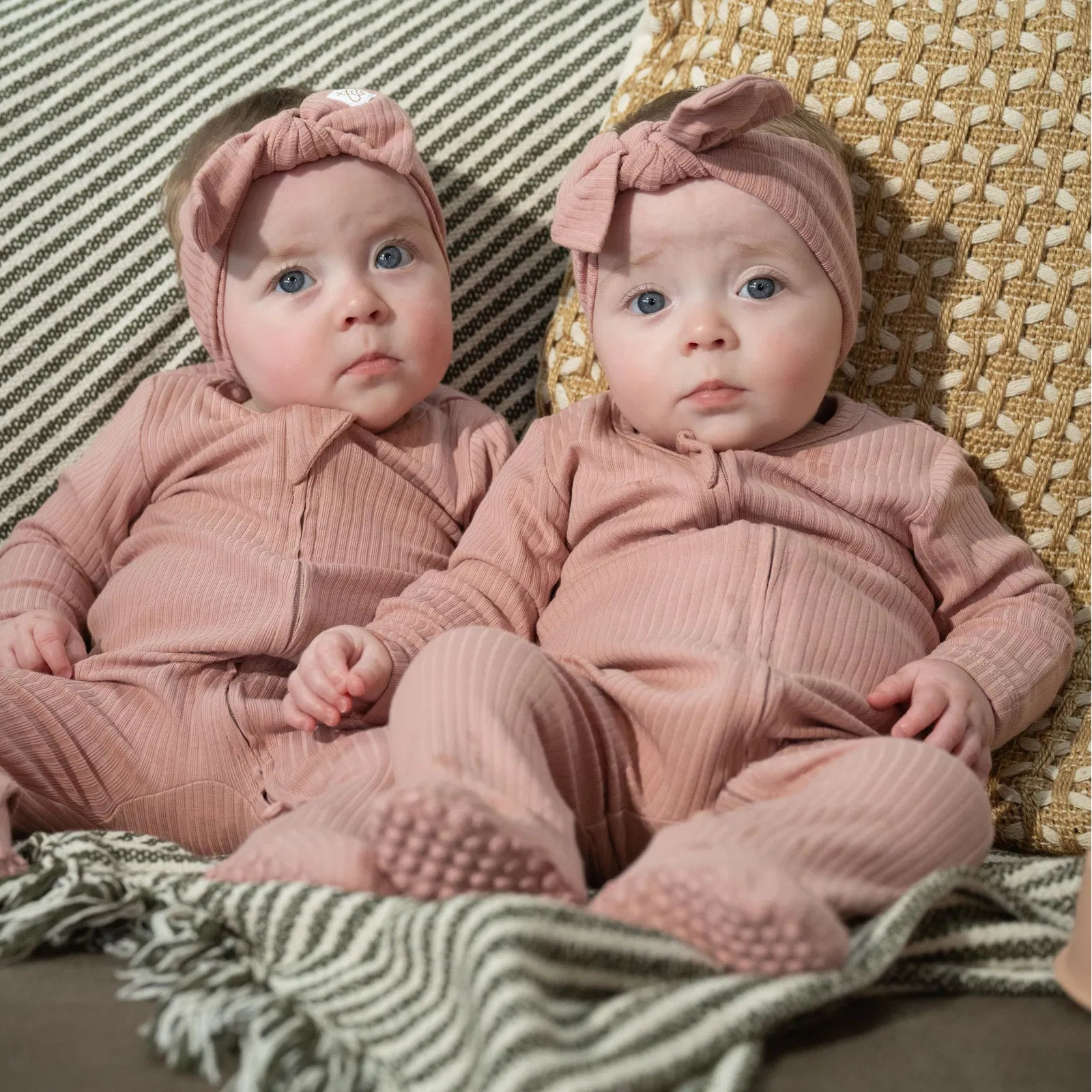 Two babies wearing The Ribbed Set Bow Edition in rose pink ribbed cotton sitting on striped and woven cushions