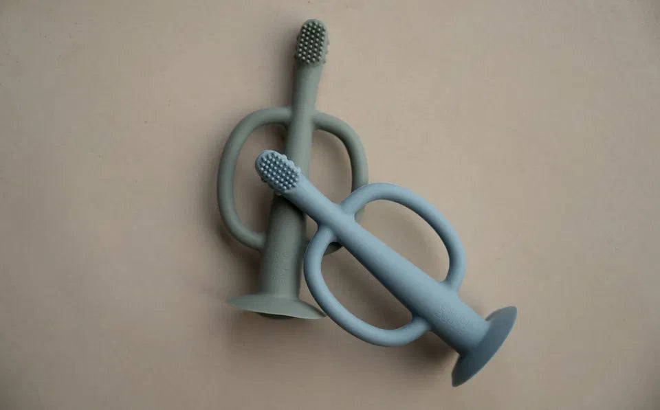 The Gentle Brush Teether baby toothbrush and teether in Stone blue and Mist Grey with soft silicone bristles and suction base.