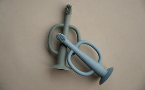The Gentle Brush Teether baby toothbrush and teether in Stone blue and Mist Grey with soft silicone bristles and suction base.