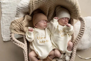 Newborn twins wearing ivory Were Here Twin Romper Set in a cozy basket with knitted hats