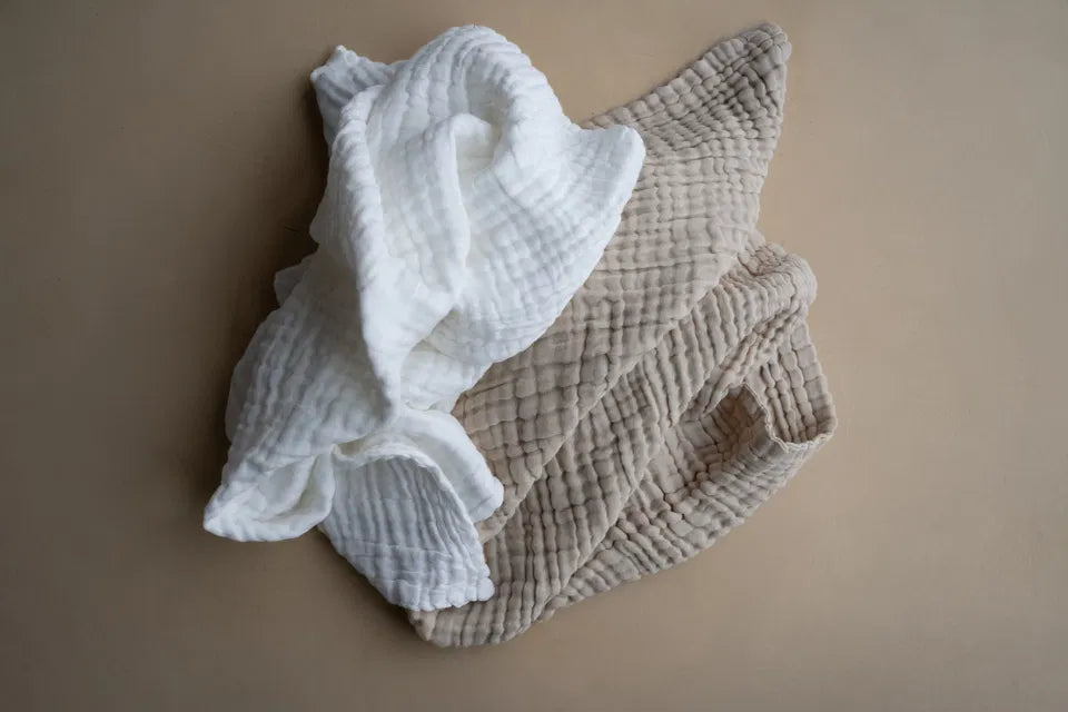 Soft breathable Snuggle Swaddle muslin blankets in Milk and Biscuit colors on beige background