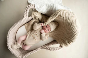 Newborn baby in biscuit beige knitted romper and hat from We’re Here twin outfit set lying in cozy basket with matching blanket
