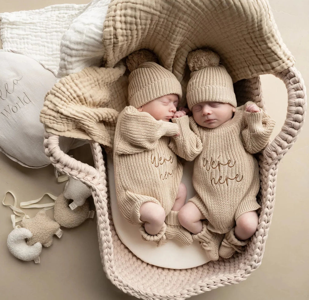 Newborn twins wearing Were Here twin outfit set biscuit beige knitted rompers with matching hats and booties in a cozy basket